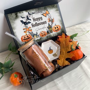 Pu&ograve; includere: Una scatola regalo nera contenente un bicchiere di vetro con coperchio in bamb&ugrave; e cannuccia, un piattino esagonale bianco con illustrazione di zucca, una collana dorata, zucche arancioni artificiali e foglie autunnali. L'interno del coperchio presenta una grafica Happy Halloween.