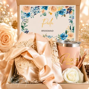 May include: A gift box containing a champagne-coloured satin robe with lace trim, a matching champagne-coloured tumbler with the name Paola and the word BRIDESMAID printed in gold, a diamond-topped pen, and a white rose, with a floral card reading Paola.