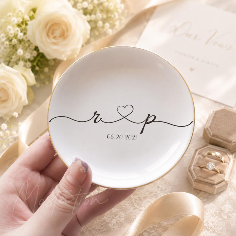 May include: A round white ceramic trinket dish with a gold-painted rim. The center features black cursive lettering with the letters r and p connected by a heart symbol, followed by the date 06.20.2021 printed in a simple black serif font.