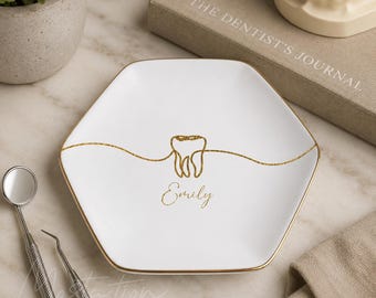 May include: A white hexagonal ceramic trinket dish with a gold-coloured rim. The centre features a gold-coloured line drawing of a tooth and the name Emily written in gold-coloured script. The dish has a smooth, glossy finish and a gold-coloured edge.