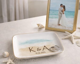 May include: A rectangular white ceramic trinket dish featuring a painted beach scene with the letters K & A and the date 10.26.2024. Two silver rings rest on the dish, which is displayed near a gold-framed photograph of a couple on a beach.