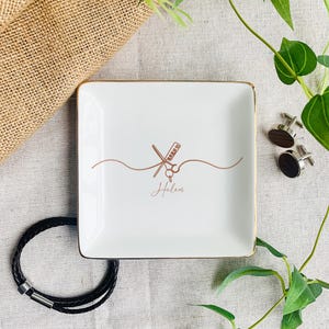 Pu&ograve; includere: Un piattino quadrato in ceramica bianca con bordo dorato. Il centro presenta un disegno a linee color rame di forbici e un pettine, con il nome Helen scritto in corsivo sotto il design, accompagnato da gemelli e un braccialetto.