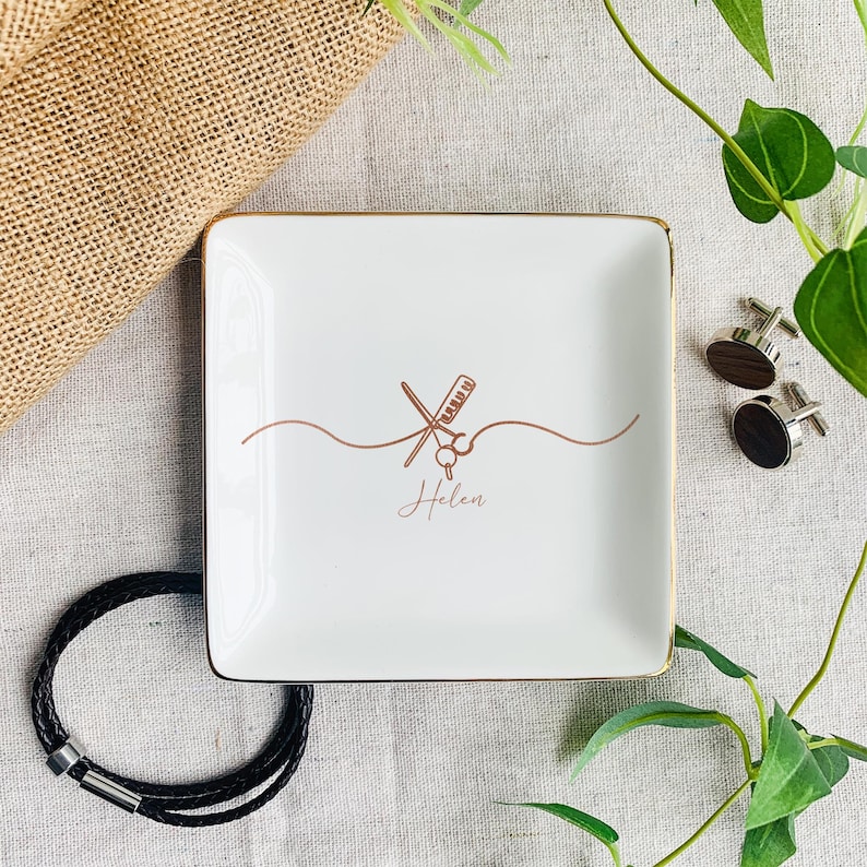 Pu&ograve; includere: Un piattino quadrato in ceramica bianca con bordo dorato. Il centro presenta un disegno a linee color rame di forbici e un pettine, con il nome Helen scritto in corsivo sotto il design, accompagnato da gemelli e un braccialetto.