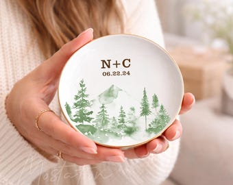May include: A round white ceramic trinket dish with a gold-toned rim. The center features a green watercolor illustration of a mountain and pine trees, with the brown text N+C and the date 06.22.24 printed above the landscape design.