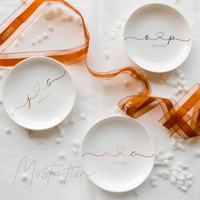 May include: Three round white ceramic trinket dishes featuring personalized script lettering and dates. One dish displays gold initials, one black, and one rose gold, each connected by a heart symbol. A sheer orange ribbon with the word HAPPY is included.
