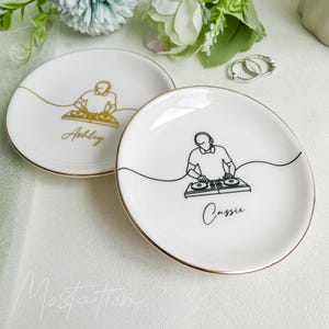 May include: Two round white ceramic trinket dishes with gold-toned rims. One dish features a gold line-art illustration of a DJ with the name Ashley, while the other features a black line-art illustration of a DJ with the name Cassie.