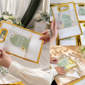 May include: A clear plastic gift bag with a gold-colored frame and handle. The bag features a sage green arch-shaped label with white floral accents and the text "Serena's Sweet 40" printed in white script. The bag is semi-transparent and rectangular.