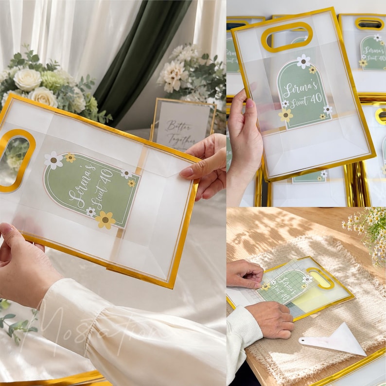 May include: A clear plastic gift bag with a gold-colored frame and handle. The bag features a sage green arch-shaped label with white floral accents and the text "Serena's Sweet 40" printed in white script. The bag is semi-transparent and rectangular.
