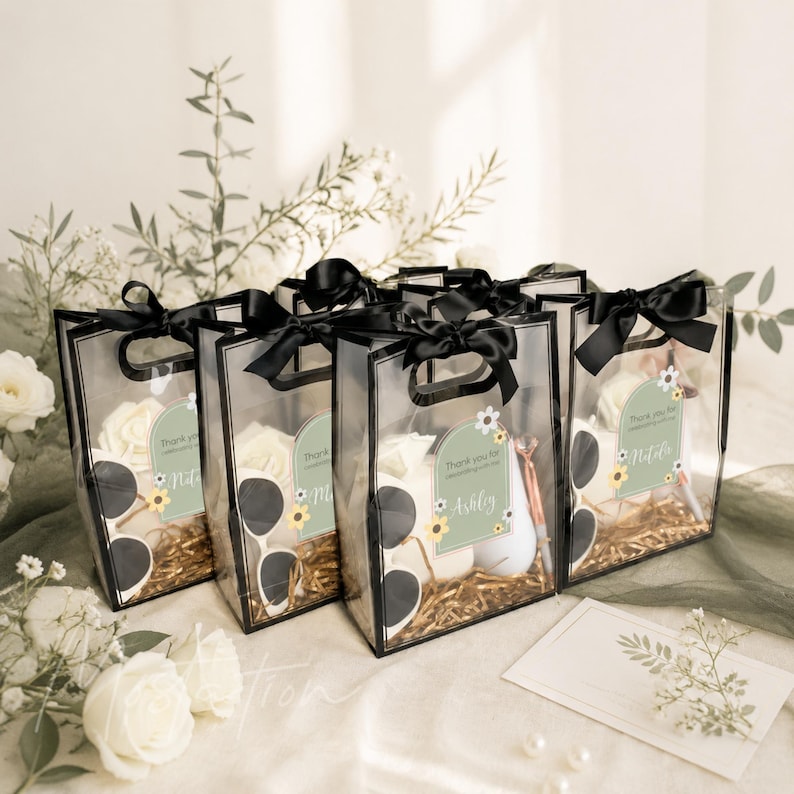 May include: Four clear rectangular gift bags with black trim and black satin ribbon bows at the top. Each bag contains white artificial flowers, a pair of white sunglasses, and a green label with the text Thank you for celebrating with us.