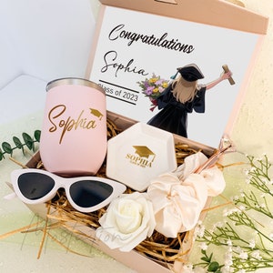 May include: A gift box containing a light pink tumbler with the name Sophia and a graduation cap icon, a white hexagonal trinket dish with gold text, white sunglasses, a white artificial rose, a cream scrunchie, and a graduation card.