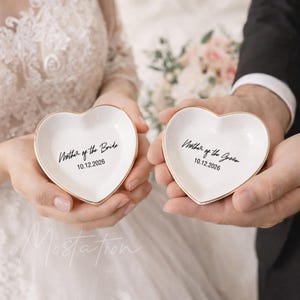 May include: Two heart-shaped white ceramic ring dishes with gold rims. One dish reads "Mother of the Bride" and the other reads "Mother of the Groom" with the date "10.12.2026" in black script. The dishes are held by two people, likely at a wedding.