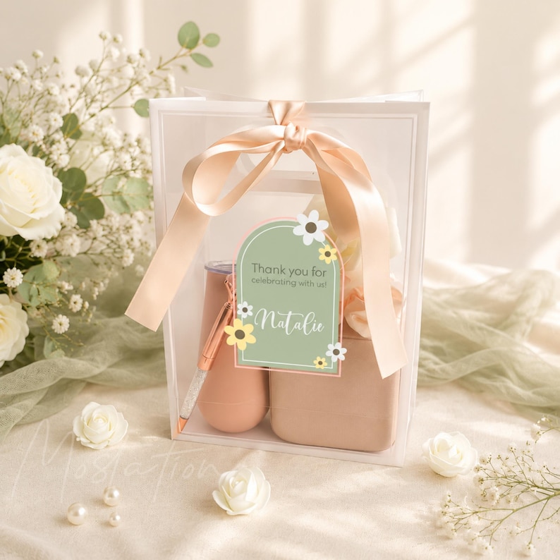 May include: A clear gift bag tied with a peach satin ribbon containing a peach-colored tumbler and a matching pen. An arched sage green tag is attached to the front, featuring white text that reads Thank you for celebrating with us! Natalie.