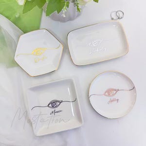 May include: Four white ceramic ring dishes with gold trim. Each dish features an eye design and a name: Dalia, Kimberly, Monica, and Ashley. The dishes are in various shapes: hexagon, rectangle, square, and circle.