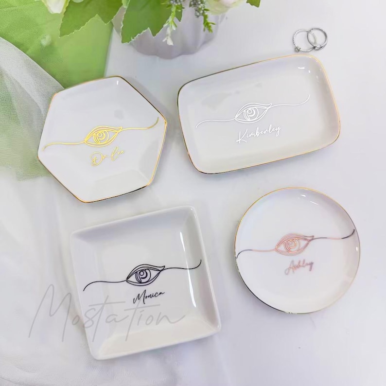 May include: Four white ceramic ring dishes with gold trim. Each dish features an eye design and a name: Dalia, Kimberly, Monica, and Ashley. The dishes are in various shapes: hexagon, rectangle, square, and circle.