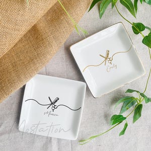 Pu&ograve; includere: Due piattini quadrati in ceramica bianca con bordi dorati. Uno presenta un disegno a linee nere di forbici e pettine con il nome Monica. L'altro presenta un disegno a linee dorate di forbici e pettine con il nome Emily.