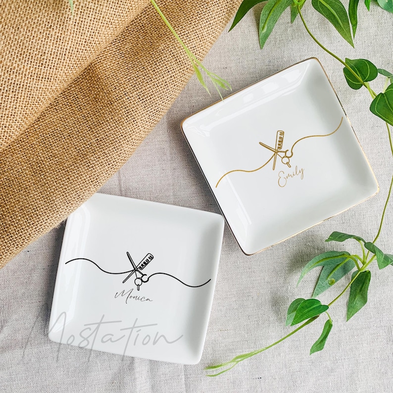 Pu&ograve; includere: Due piattini quadrati in ceramica bianca con bordi dorati. Uno presenta un disegno a linee nere di forbici e pettine con il nome Monica. L'altro presenta un disegno a linee dorate di forbici e pettine con il nome Emily.