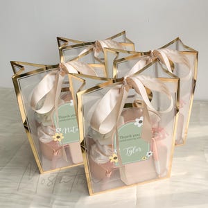 May include: Four clear gift bags with gold metallic trim and beige satin ribbon bows. Each bag contains a pink pouch and a tag with the text Thank you celebrating with us and a name, decorated with small white and yellow flower illustrations.