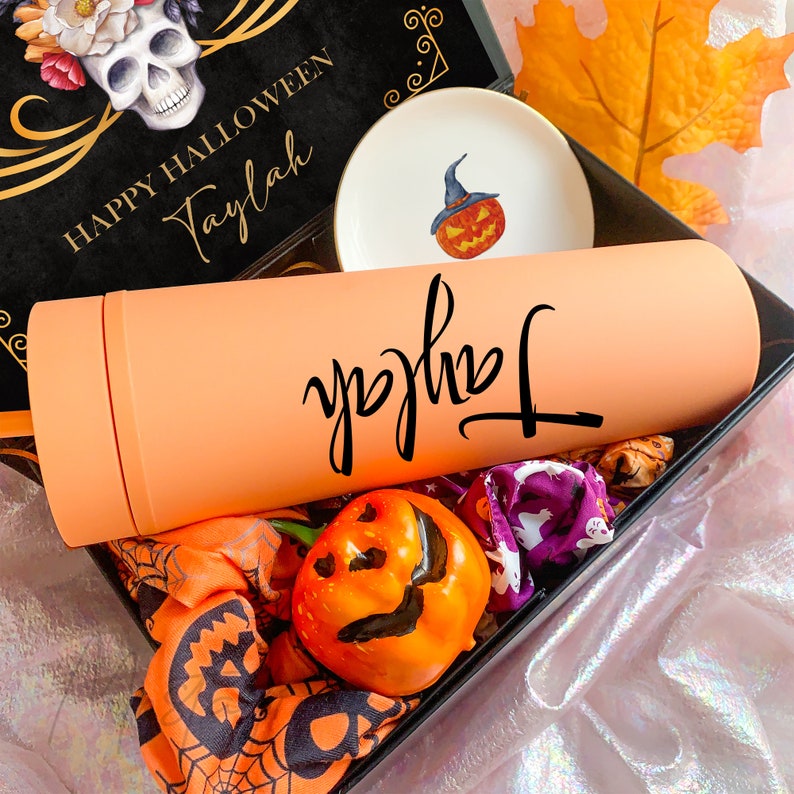 May include: A gift box containing a matte peach tumbler with the name Taylah in black script, a small white dish with a pumpkin illustration, a miniature orange pumpkin figurine, and fabric featuring ghost and spiderweb patterns.