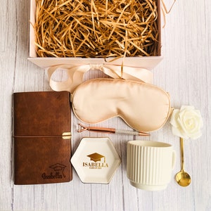 May include: A graduation gift box with a brown leather journal, sleep mask, pen, ring dish, mug, and spoon. The ring dish is white with gold text that reads "Isabella Class of 2024". The box is pink and filled with straw.