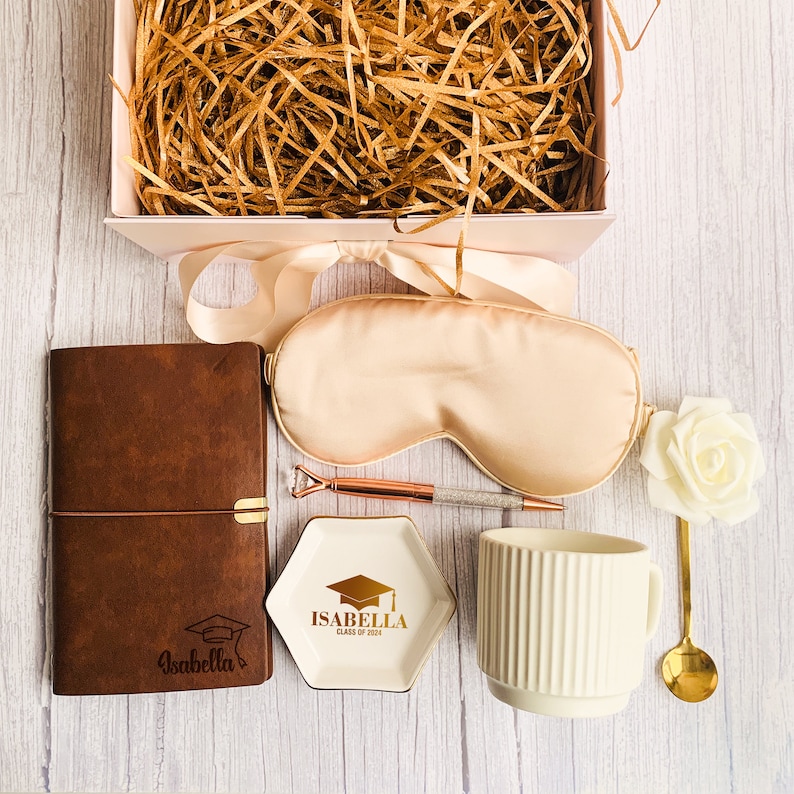 May include: A graduation gift box with a brown leather journal, sleep mask, pen, ring dish, mug, and spoon. The ring dish is white with gold text that reads "Isabella Class of 2024". The box is pink and filled with straw.