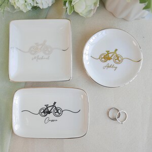 May include: Three white ceramic trinket dishes with gold-trimmed edges. One square dish features a silver bicycle graphic and the name Michael. One round dish has a gold bicycle graphic and the name Ashley. One rectangular dish has a black bicycle graphic and the name Cassie.