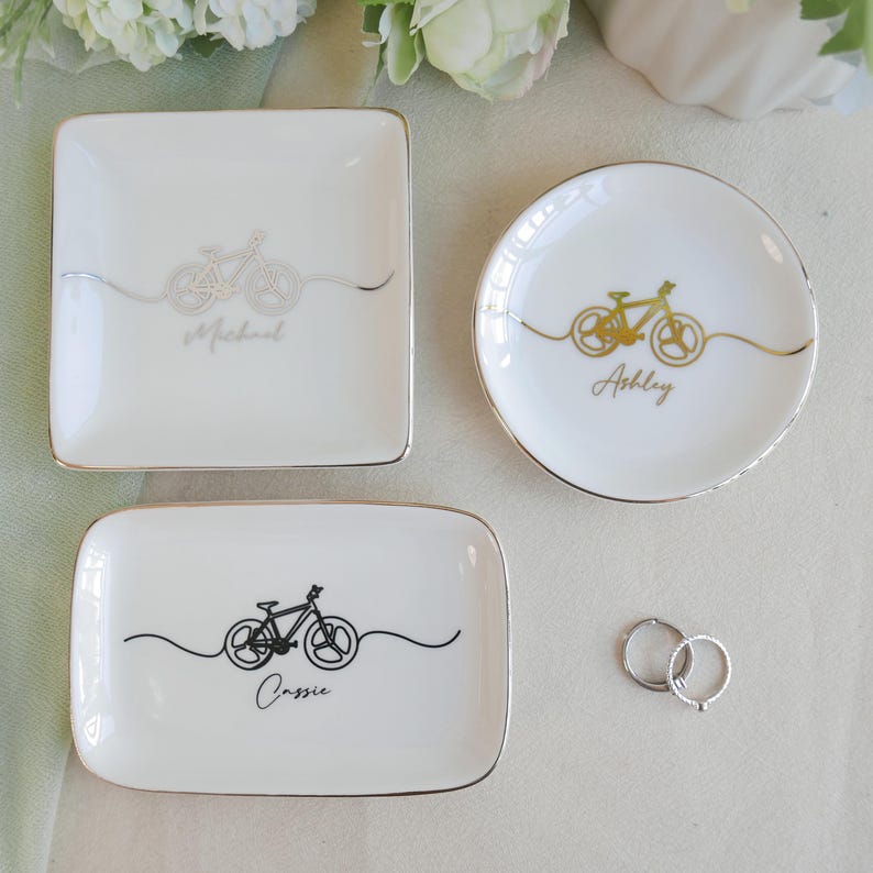 May include: Three white ceramic trinket dishes with gold-trimmed edges. One square dish features a silver bicycle graphic and the name Michael. One round dish has a gold bicycle graphic and the name Ashley. One rectangular dish has a black bicycle graphic and the name Cassie.