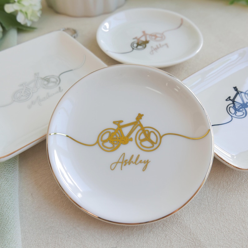 May include: A collection of white ceramic trinket dishes with gold-rimmed edges. Each dish features a continuous line drawing of a bicycle in gold, silver, or dark blue ink. The name Ashley is printed in gold cursive script on the central round dish.