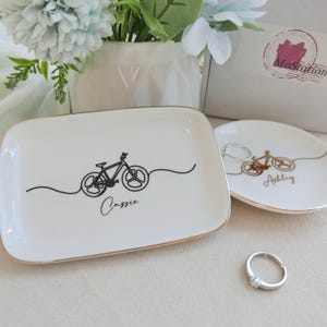 May include: A rectangular white ceramic trinket tray with a black line art bicycle and the name Cassie, alongside a round white ceramic trinket tray with a gold line art bicycle and the name Ashley, both featuring gold-toned rims.