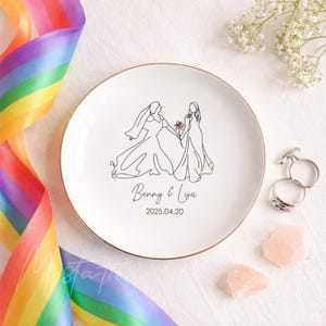 Może przedstawiać: Biały ceramiczny talerz ze złotym obrzeżem z czarną grafiką liniową przedstawiającą dwie postacie w sukniach ślubnych trzymające mały kolorowy kwiat. Tekst Benny & Lisa oraz data 2025.04.20 są nadrukowane czarnym kolorem pod ilustracją.
