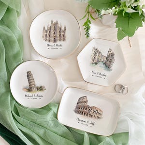 May include: Four white ceramic ring dishes with gold trim. Each dish features a different iconic Italian landmark: the Duomo in Milan, the Rialto Bridge in Venice, the Leaning Tower of Pisa, and the Colosseum in Rome. Each dish has a personalized inscription with a couple's names and wedding date.
