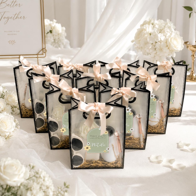 May include: A collection of clear gift bags with black trim, each tied with a pale pink ribbon. Inside each bag are white sunglasses, a white bottle, a rose-shaped item, and a pen. A green tag on each bag reads Thank you for celebrating with us Natalie.