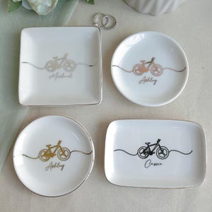 May include: Four white ceramic trinket dishes in square, round, and rectangular shapes with metallic rims. Each dish features a minimalist line-art bicycle graphic and a personalised name in script: Michael, Ashley, or Cassie in gold or black ink.