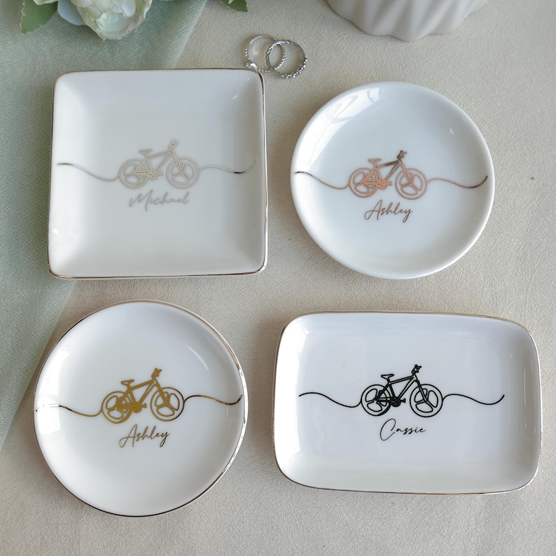 May include: Four white ceramic trinket dishes in square, round, and rectangular shapes with metallic rims. Each dish features a minimalist line-art bicycle graphic and a personalised name in script: Michael, Ashley, or Cassie in gold or black ink.