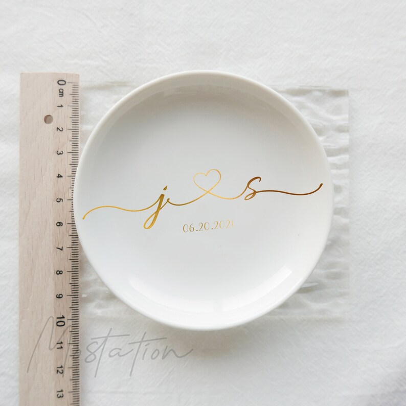 May include: A round white ceramic trinket dish featuring gold calligraphy of the letters j and s connected by a heart, with the date 06.20.2021 printed below. A wooden ruler placed alongside shows the dish has a diameter of approximately 4 inches.