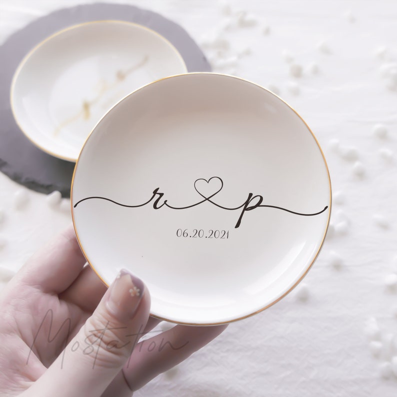 May include: A round white ceramic trinket dish with a gold-painted rim. The center features black cursive lettering of the letters r and p connected by a heart symbol, with the date 06.20.2021 printed in a simple black font underneath the letters.