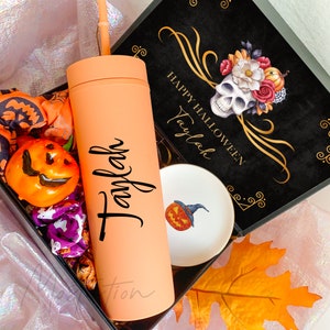 May include: A matte peach-colored cylindrical tumbler with a matching straw, featuring the name Taylah in black script. It is presented in a black gift box with a skull and floral graphic, a small white dish with a pumpkin, and orange fabric.