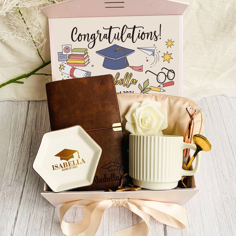May include: A graduation gift box with a pink lid and a cream-colored ribbon. Inside, a brown leather journal, a white mug, a small dish with gold lettering, and a cosmetic bag. The box lid says "Congratulations!" and "Isabella Class of 2024."