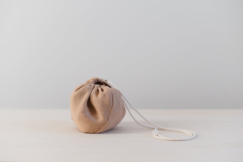 May include: A light brown drawstring bag with a white cord. The bag is sitting on a light wood surface.