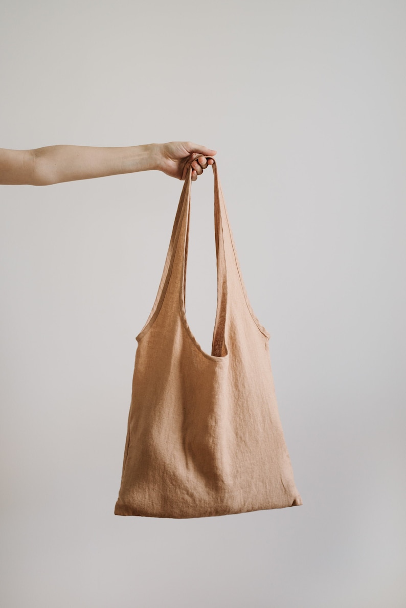 May include: A light brown linen tote bag with a single handle. The bag is empty and hanging from a hand.