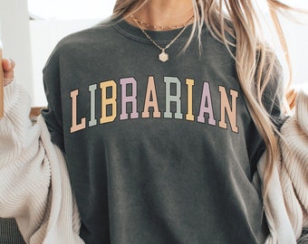 May include: A woman wearing a gray t-shirt with the word "LIBRARIAN" printed in rainbow colors. She is sitting on a white fluffy chair with a cup of coffee in her hand.