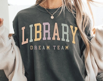 May include: Dark grey sweatshirt with the text "LIBRARY DREAM TEAM" in pastel colors. The sweatshirt is worn with shorts and a cream-colored cardigan. A glass of iced coffee is held in the hand.