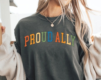 May include: A woman wearing a grey t-shirt with the text "PROUD ALLY" in rainbow colours. The shirt is a relaxed fit and has a crew neck.