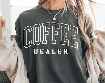 Könnte beinhalten: Ein dunkelgraues Sweatshirt mit dem Text "COFFEE DEALER" in weißen Blockbuchstaben. Das Sweatshirt wird mit Shorts und einer cremefarbenen Strickjacke getragen. Ein Glas Eiskaffee wird in der Hand gehalten.