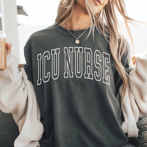 May include: A woman wearing a dark gray t-shirt with the text "ICU NURSE" in white outlined letters. She is sitting on a white furry chair with a cup of coffee in her hand.
