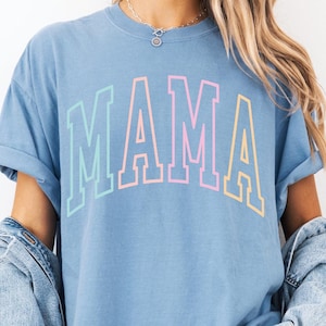 May include: A light blue t-shirt with the word "MAMA" printed in rainbow colors. The shirt has a relaxed fit and rolled up sleeves.