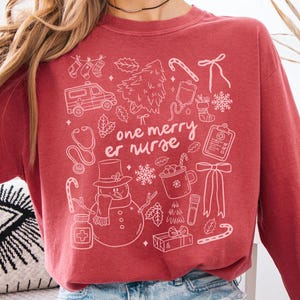 May include: Crimson-colored sweatshirt with a white graphic design featuring Christmas-themed illustrations and the text "one merry er nurse." The design includes a snowman, candy canes, and medical symbols. The sweatshirt is worn with denim shorts.