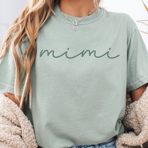 May include: A light green t-shirt with the word "mimi" written in cursive on the front. The shirt is being worn by a person with long blonde hair and is paired with denim shorts.