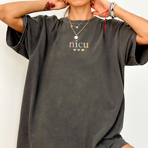 May include: A dark gray t-shirt with the word "NICU" embroidered in a soft, pastel color palette, with three small hearts below. The shirt has a relaxed fit and short sleeves. The word "PEPPER" is printed at the bottom left.