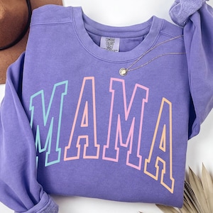May include: A purple sweatshirt with the word "MAMA" printed in rainbow colors. The sweatshirt has a crew neck and long sleeves.
