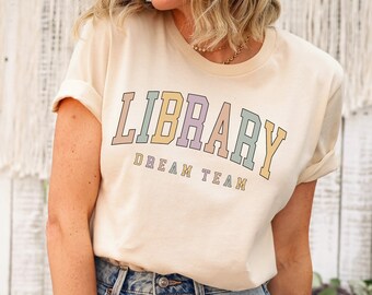 May include: A cream colored t-shirt with the text "LIBRARY DREAM TEAM" in rainbow colors. The shirt is being worn by a person with light brown hair and blue jeans.